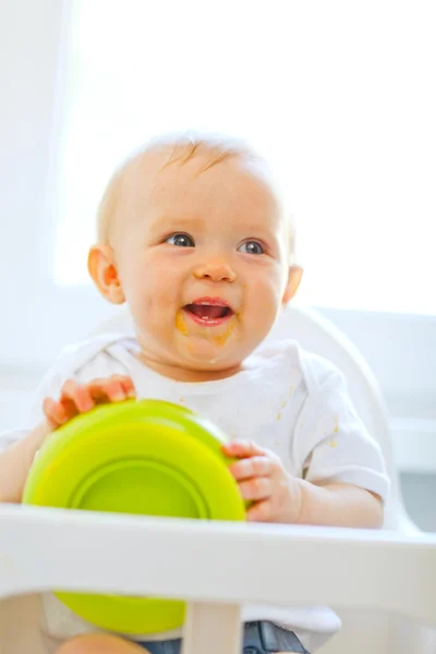 Eat smeared cheerful baby girl in baby chair playing with plate — Stock Photo, Image