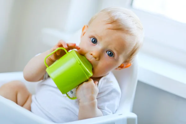 Preciosa niña sentada en la silla y bebiendo de la taza del bebé — Foto de Stock