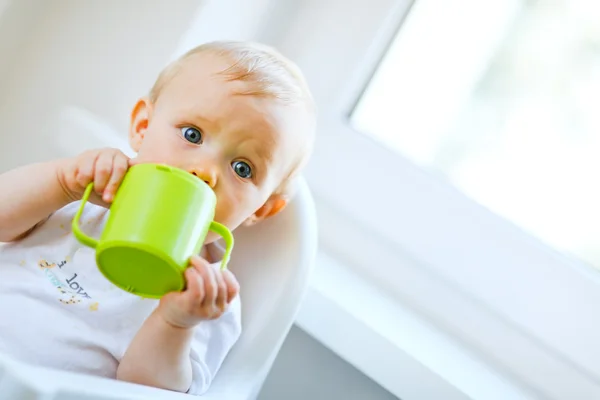 Bonita niña sentada en la silla y bebiendo de la taza del bebé — Foto de Stock