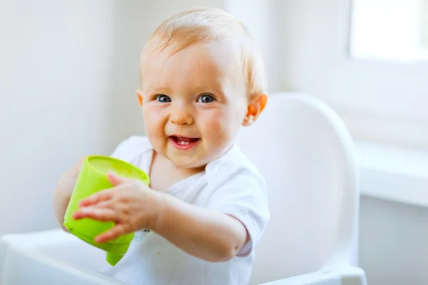 Cheerful baby girl sitting in chair and holding baby cup — Stock Photo, Image