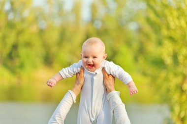 Mother's hands holding laughing adorable baby against sunny leaves clipart