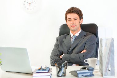 Smiling modern businessman with crossed arms on chest sitting at office des clipart