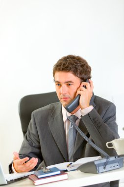 Serious modern businessman sitting at office desk and speaking p clipart