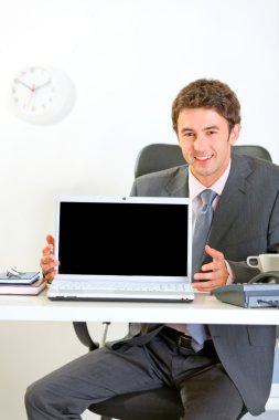 Smiling businessman sitting at office desk and showing laptops b clipart