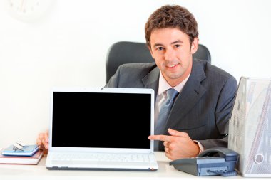 Smiling businessman sitting at office desk and pointing finger on laptop wi clipart