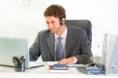 Smiling modern manager with headset looking in laptop clipart