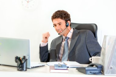 Smiling businessman with headset sitting at office desk and looking on lap clipart