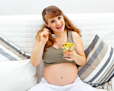 Smiling beautiful pregnant woman sitting on sofa with fruit salad in hand clipart