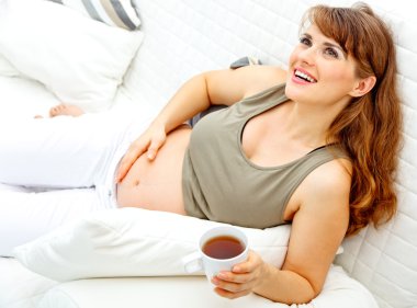 Smiling beautiful pregnant woman relaxing on couch and holding cup of tea clipart