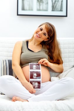 Smiling beautiful pregnant woman sitting on sofa with echo in hand. clipart