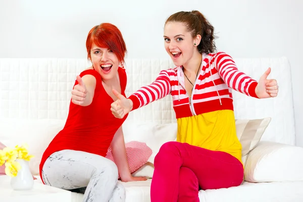 Two smiling girlfriends sitting on sofa and showing thumbs up gesture — Stock Photo, Image