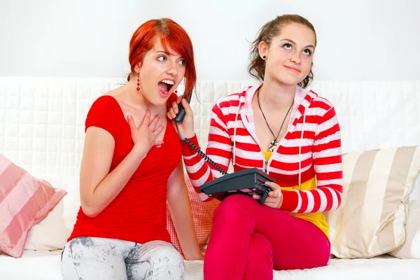 Funny girl sitting on sofa and holding phone at ear her surprised girlfrien — Stock Photo, Image