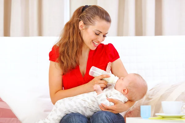 Happy mother sitting on sofa and feeding baby — Stock Photo, Image