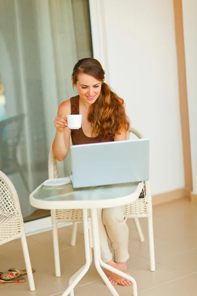 Mujer bonita sonriente tomando una taza de té y mirando en el portátil —  Fotos de Stock