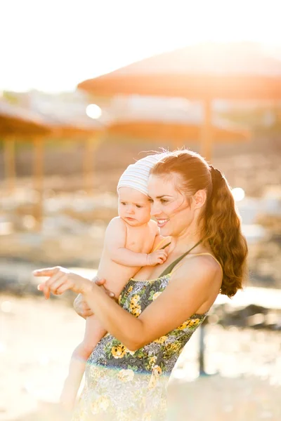 Portrait of young mother with baby on beach showing something by — Stockfoto