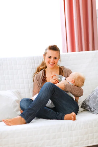 Young mother sitting on sofa and feeding her baby — Stock Photo, Image