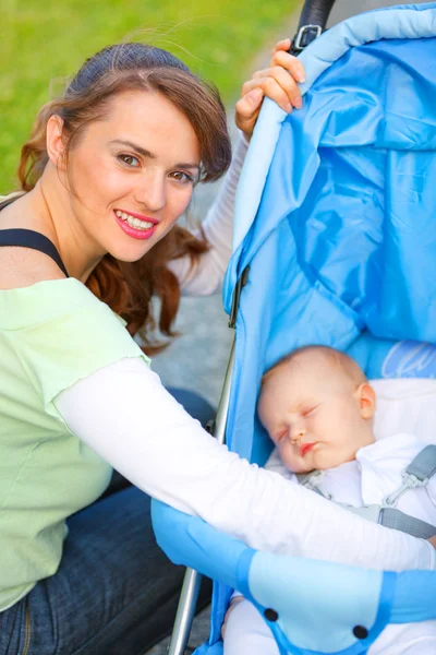 Lachende moeder zitten in de buurt van wandelwagen en verzorgen over haar baby — Stockfoto