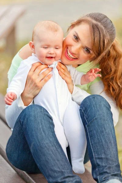 Happy mother sitting on bench in park and holding smiling adorable baby in — Stock Photo, Image