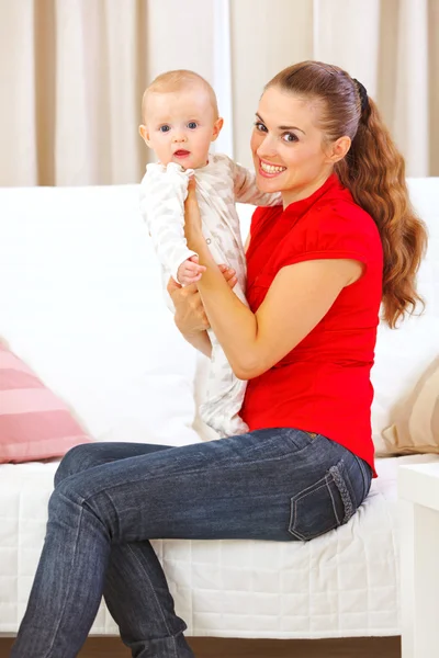 Happy mother sitting on divan and holding adorable baby in hands — Stock Photo, Image