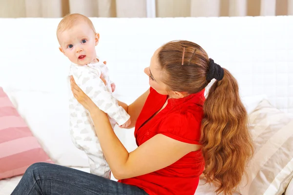 Mor sitter på divan och håller intersted baby i händer — Stockfoto