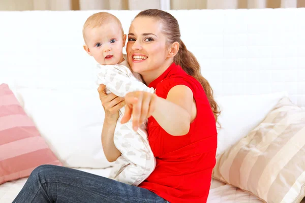 Mãe feliz sentada no divã e mostrando algo para seu bebê — Fotografia de Stock