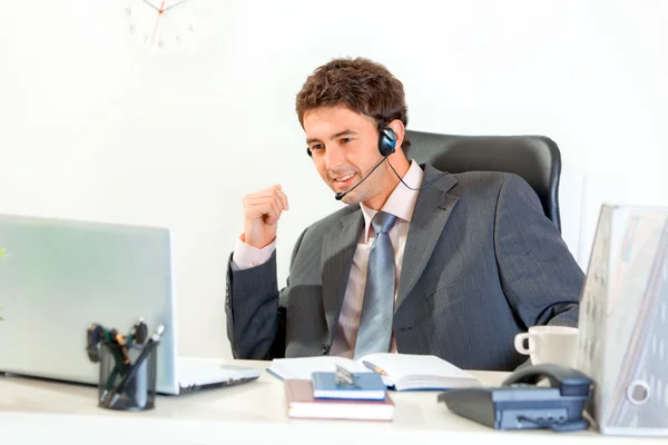 Hombre de negocios sonriente con auriculares sentado en el escritorio de la oficina y mirando en el regazo — Foto de Stock