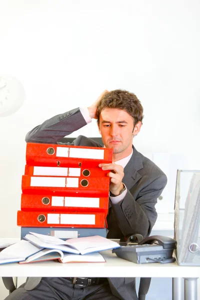 Confused businessman sitting at office desk with pile of folders — Stock Photo, Image