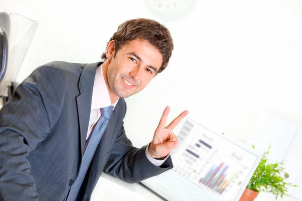 Hombre de negocios sonriente sentado en el escritorio de la oficina con el ordenador portátil y mostrando la victoria —  Fotos de Stock