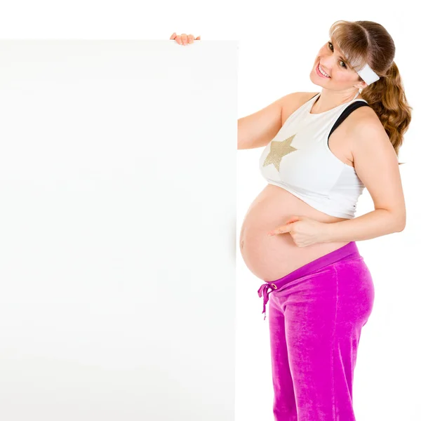 Улыбающаяся красивая беременная женщина, указывающая пальцем на чистый рекламный щит — стоковое фото