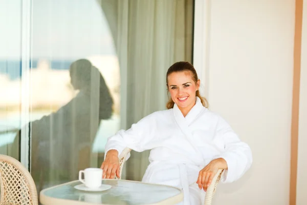 Lächelnde hübsche Frau im Bademantel sitzt am Tisch auf der Terrasse — Stockfoto