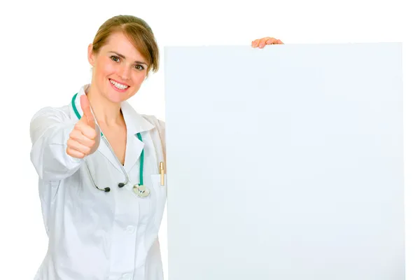 Médico sonriente mujer médico sosteniendo cartelera en blanco y mostrando los pulgares hacia arriba —  Fotos de Stock