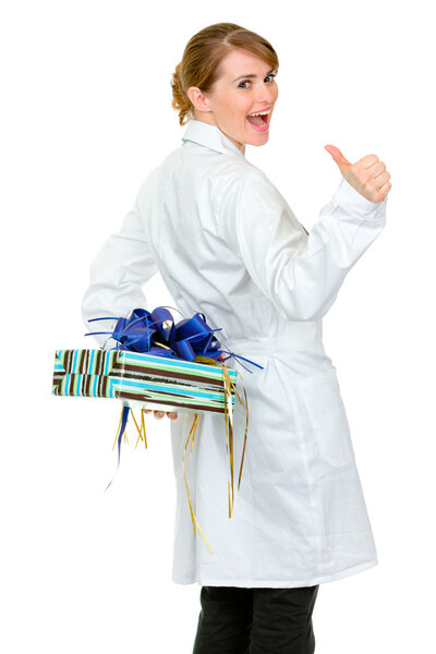 Happy medical doctor woman holding gift behind her back and thumbs up