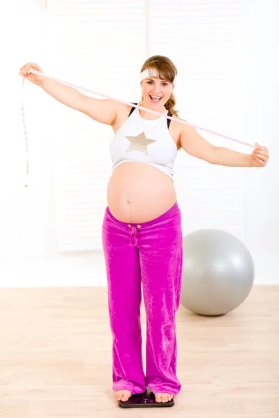 Mulher grávida sorrindo em pé na balança de peso e segurando fita métrica — Fotografia de Stock
