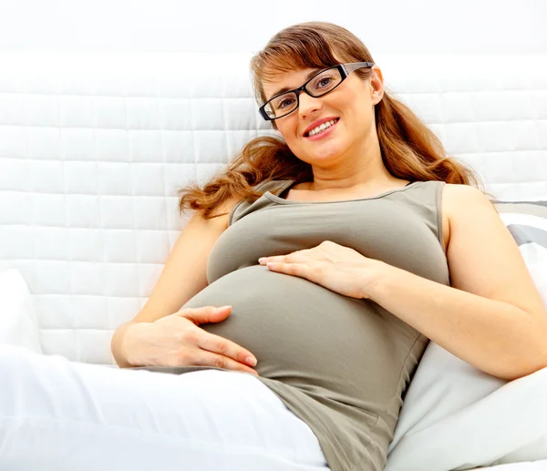 Lächelnde schöne schwangere Frau, die auf dem Sofa liegt und ihren Bauch hält. — Stockfoto