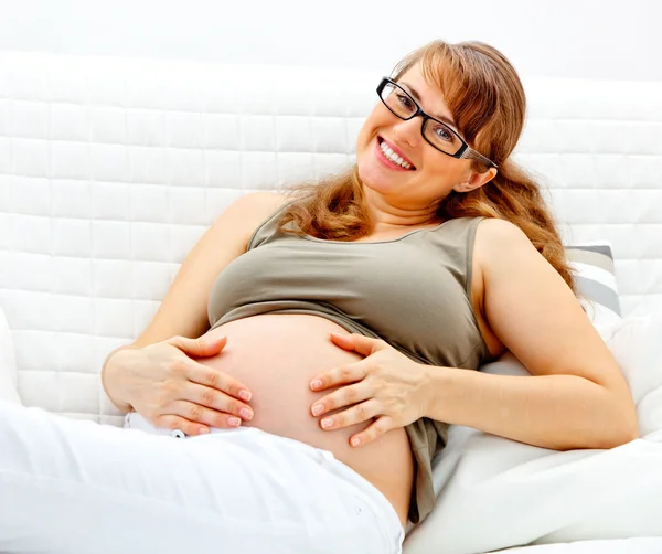 Sonriendo hermosa mujer embarazada sentada en el sofá y sosteniendo su vientre — Foto de Stock