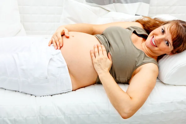 Szép terhes nő pihentető a kanapén, és ő poci gazdaság mosolyogva. — Stock Fotó