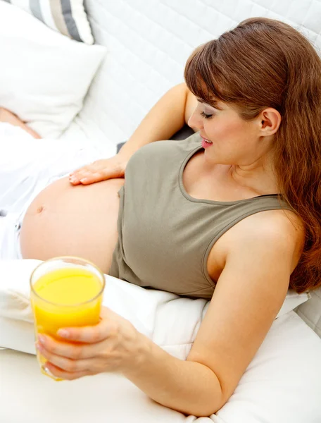 Pięknych kobiet w ciąży, relaksując się na kanapie z szklanką soku w ręku — Zdjęcie stockowe