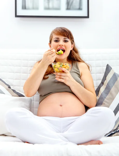 Ευτυχής όμορφη έγκυος γυναίκα κάθεται στον καναπέ και να τρώνε φρούτα σαλάτα — Φωτογραφία Αρχείου