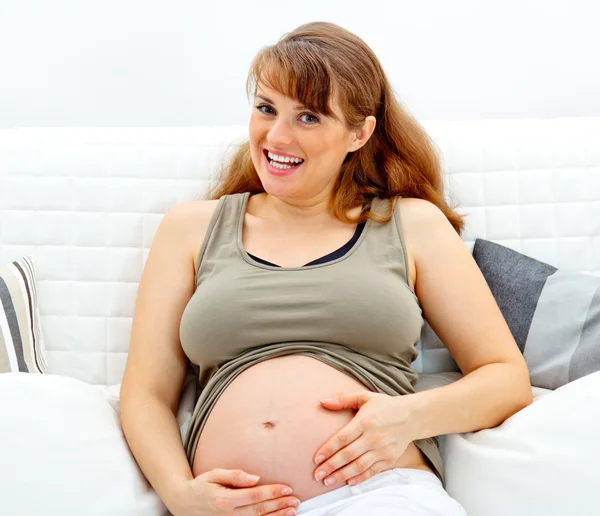 Sonriendo hermosa embarazada sentada en el sofá y tocando su vientre —  Fotos de Stock
