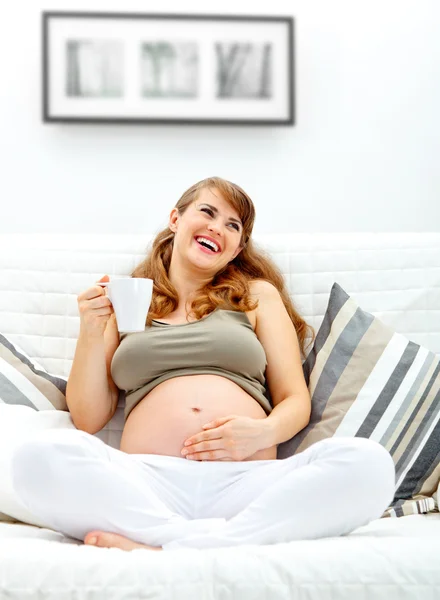 Rindo bela mulher grávida relaxando no sofá e segurando xícara de chá — Fotografia de Stock