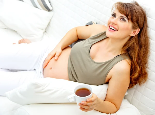 Kanepede rahatlatıcı ve çay tutan güzel bir hamile kadın gülümsüyor — Stok fotoğraf