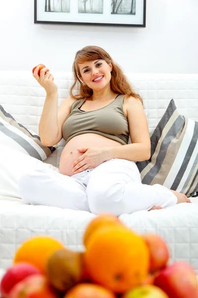 Divanda evde oturan ve meyve tutan güzel bir hamile kadın — Stok fotoğraf