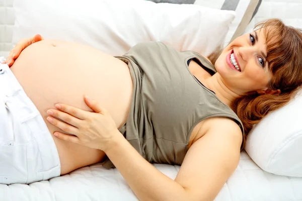 Sorrindo bela fêmea grávida deitada no sofá e segurando sua barriga . — Fotografia de Stock