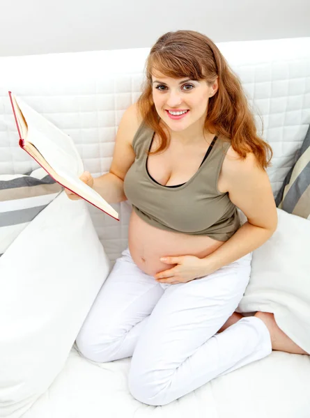 Sonriendo hermosa mujer embarazada sentada en un sofá con libro . — Foto de Stock