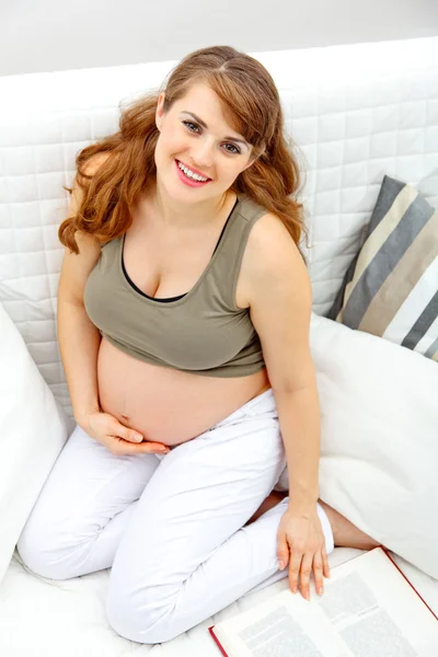도 서와 함께 소파에 앉아 웃는 아름 다운 임신한 여자 — 스톡 사진