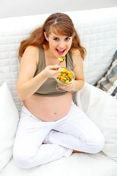 Усміхнена красива вагітна жінка сидить на дивані і їсть фруктовий салат — стокове фото