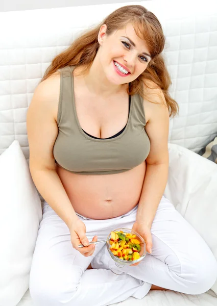 Happy beautiful pregnant woman sitting on sofa with fruit salad in hand. — Stock Photo, Image