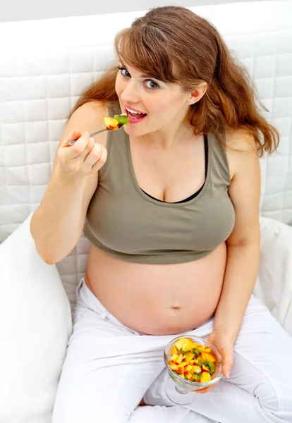 Smiling beautiful pregnant woman sitting on sofa and eating fruit salad — Stok fotoğraf