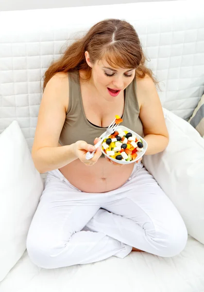 Усміхнена красива вагітна жінка сидить на дивані і їсть свіжий овоч — стокове фото