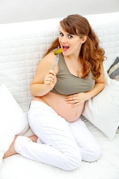 Sonriendo hermosa mujer embarazada sentada en el sofá y comiendo pepinillos —  Fotos de Stock